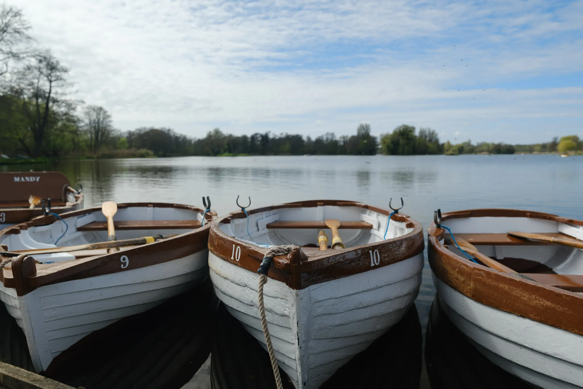 three small boats tied to a dock on a lake with tress and a cloudy, blue sky in the background.