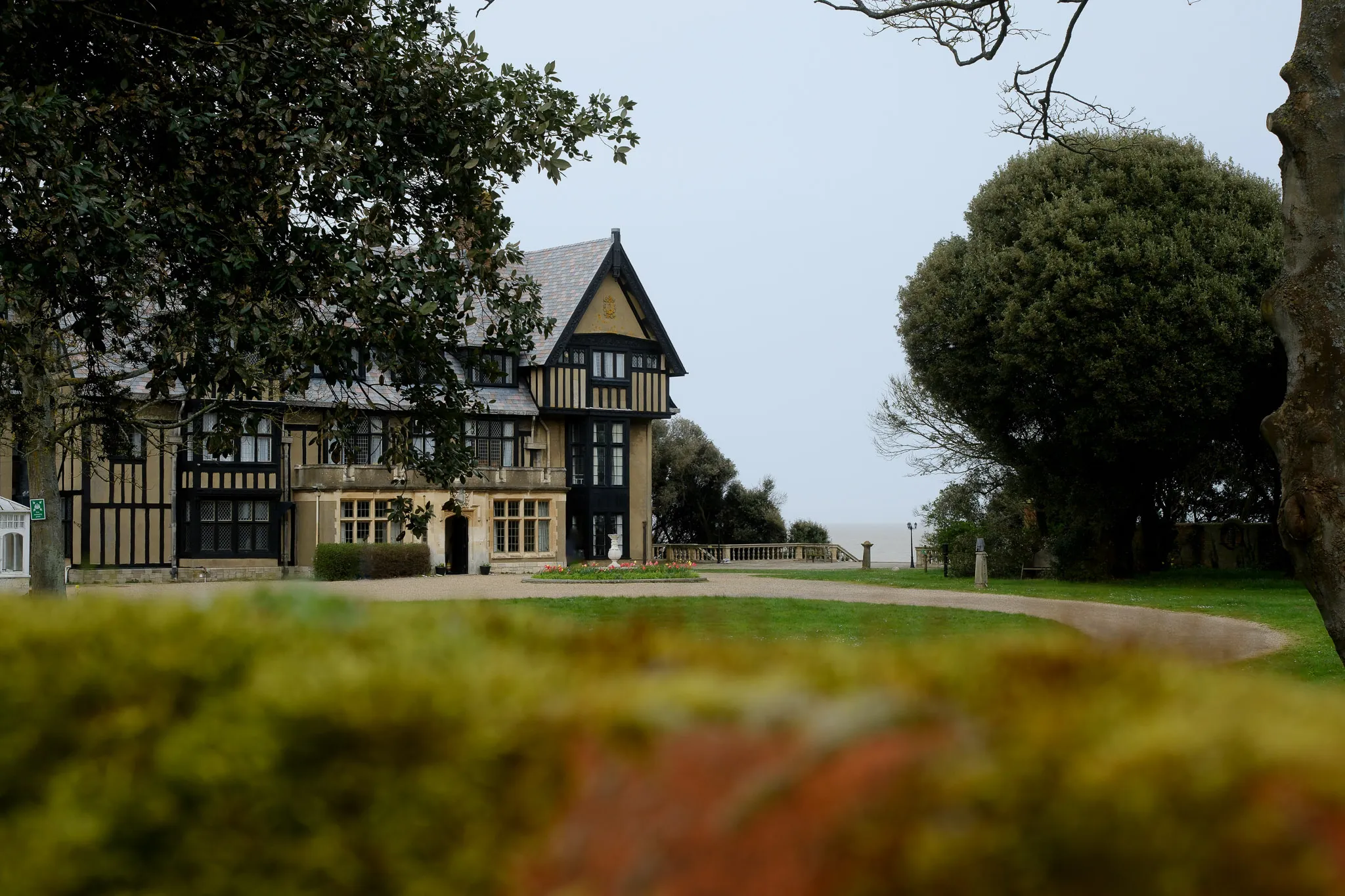 a large manor house sitting on top of a drive, surrounded by well-kept grass and a massive tree.