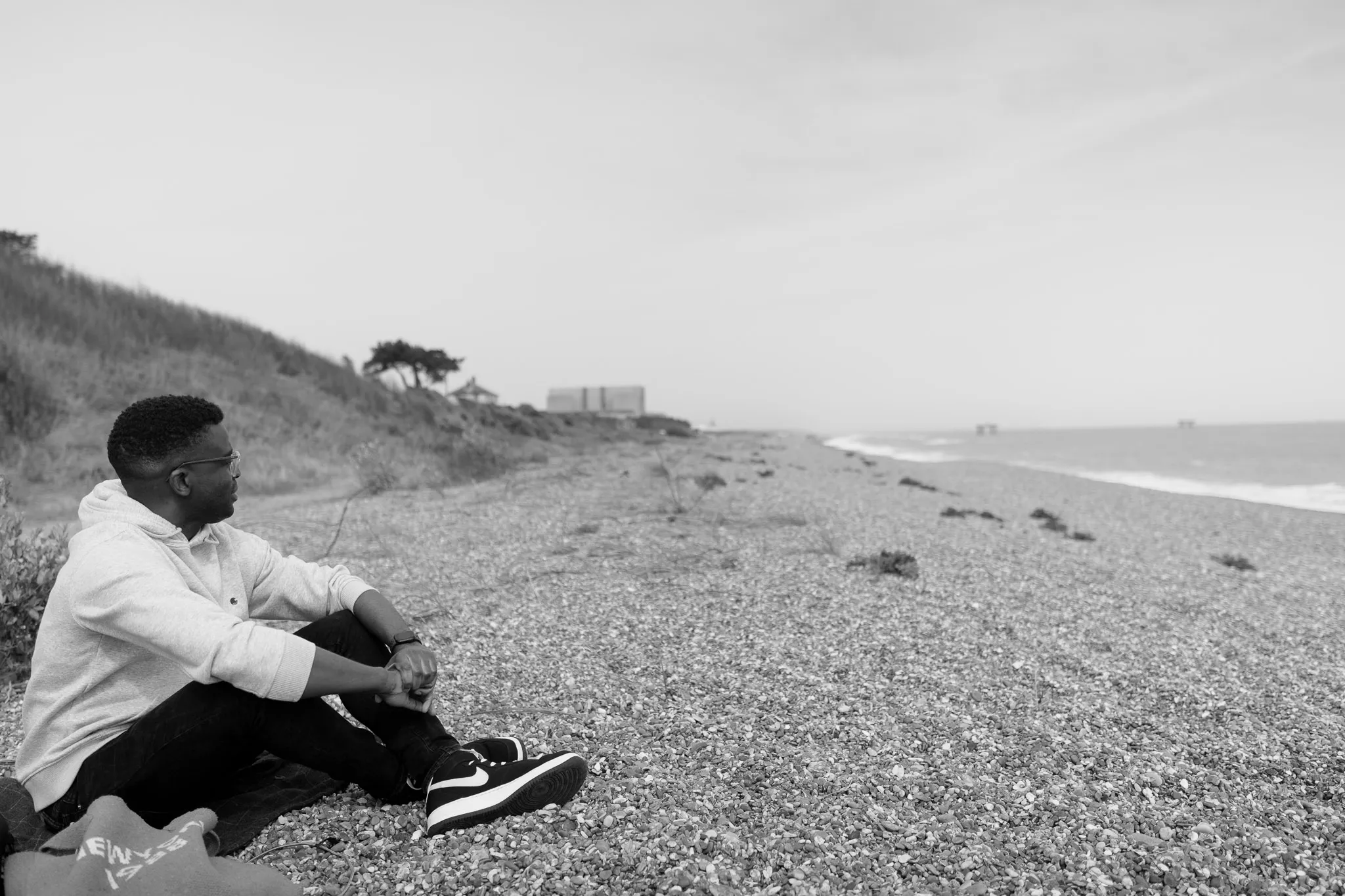 man sat down on a stoney beach with legs crossed looking away the camera, deep in thought.