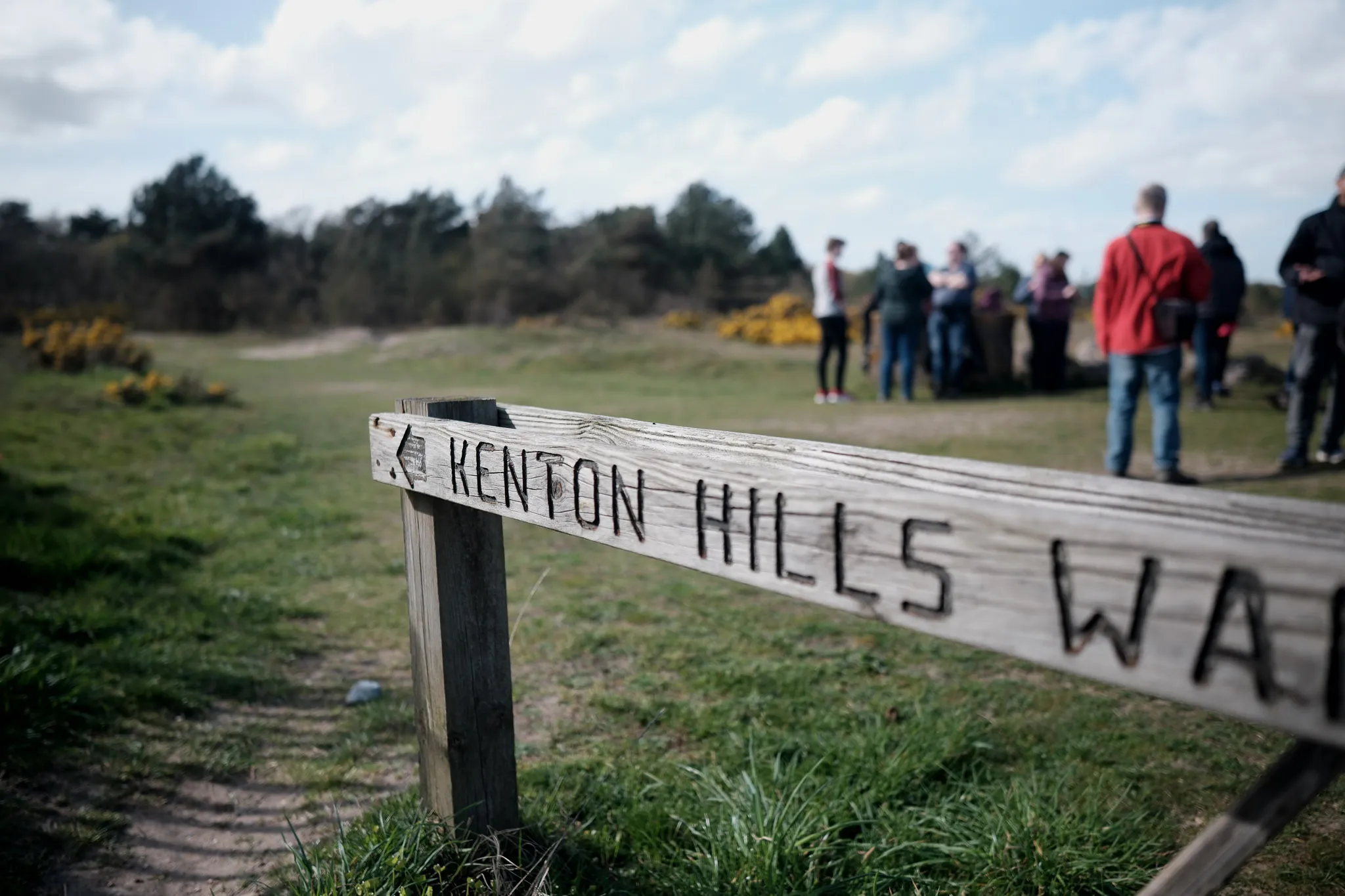 group of people standing behind an in-focus, slighty worn, wooden sign pointing towards "Kenton Hills..."